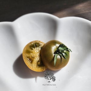 Op de afbeelding: Een gehalveerde groene tomaat met een gele binnenkant en een hele groene tomaat met een groene steel op een wit bord. Het bord heeft een gegolfde rand. De tekst "Head ToMaToes Seeds" is op het bord gedrukt.