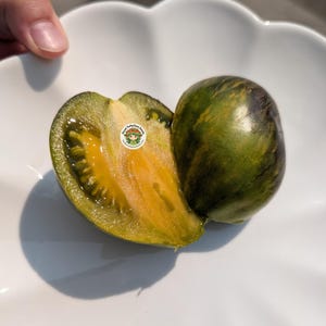 Può includere: Un pomodoro tagliato con buccia a strisce verde scuro e viola e una polpa interna giallo oro vibrante. Un piccolo adesivo circolare con il testo Head ToMeToes Seeds e un personaggio dei cartoni animati è posizionato sulla superficie tagliata.