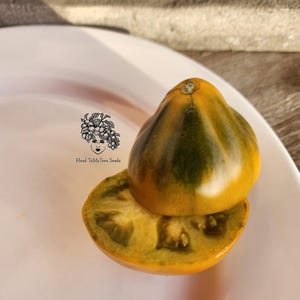 May include: A halved heirloom tomato with yellow and green stripes, resting on a white plate. The cut side reveals the tomato's interior. The image includes the text "Head ToMaToes Seeds" with a graphic of a woman's face.