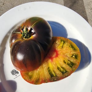 Puede incluir: Primer plano de un tomate de herencia de color púrpura oscuro y verde, con un corte que revela un interior amarillo y rojo. El tomate está sobre un plato blanco, mostrando su color y textura únicos. La imagen está bien iluminada.