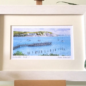 May include: A framed watercolor painting of a pier extending into the water with a white cliff in the background. The painting is titled "Sandgate Pier" and signed "Robin Downie".