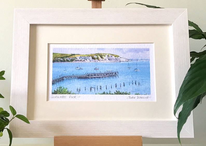 May include: A framed watercolor painting of a pier extending into the water with a white cliff in the background. The painting is titled "Sandgate Pier" and signed "Robin Downie".