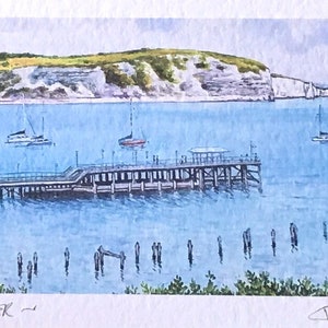 May include: A watercolor painting of a pier extending out into a body of water with white cliffs in the background. The pier has a wooden structure and is surrounded by blue water. The painting is titled "Swanage Pier" and signed "John Downie".