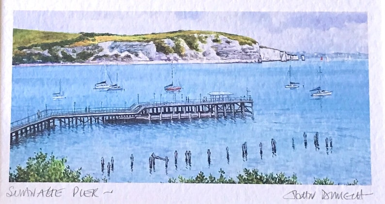 May include: A watercolor painting of a pier extending out into a body of water with white cliffs in the background. The pier has a wooden structure and is surrounded by blue water. The painting is titled "Swanage Pier" and signed "John Downie".