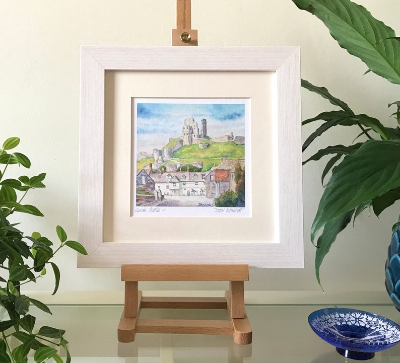 May include: A framed watercolor painting of a castle on a hill, with a village in the foreground. The painting is in a white frame and displayed on a small wooden easel. The painting is titled "Corfe Castle".
