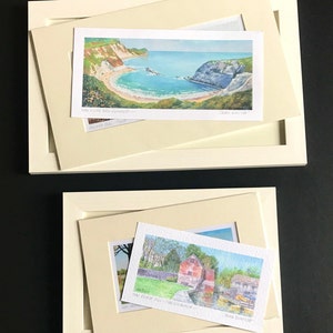 May include: Two framed watercolor paintings. The top painting shows a scenic view of a bay with a beach and cliffs. The bottom painting shows a red brick building with a river and boats. The top painting is titled "Lulworth Cove" and the bottom painting is titled "The Black Mill - Hamsterley".