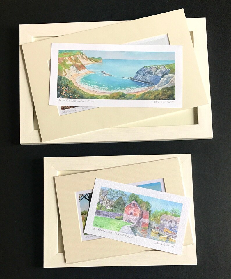 May include: Two framed watercolor paintings. The top painting shows a scenic view of a bay with a beach and cliffs. The bottom painting shows a red brick building with a river and boats. The top painting is titled "Lulworth Cove" and the bottom painting is titled "The Black Mill - Hamsterley".