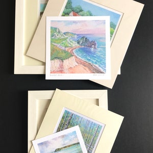 May include: Several framed watercolor landscape paintings. One features a coastal scene with a beach and cliffs, another depicts a forest of birch trees, and a third shows a body of water. The frames are off-white.