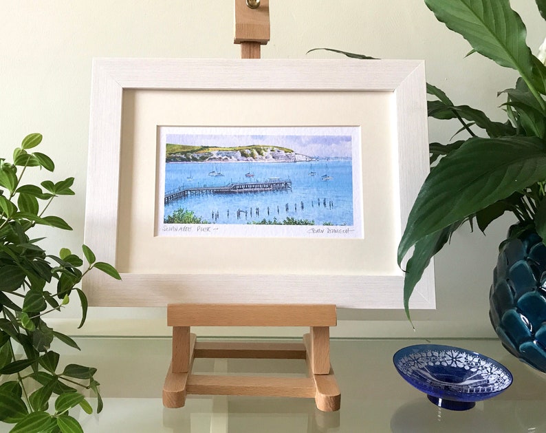 May include: A framed watercolor painting of a pier with boats in the water. The painting is titled "Sandbanks Pier" and signed "Dave Somerset".