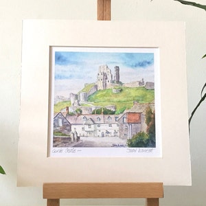 May include: Watercolor painting of Corfe Castle, set on a green hill under a blue sky. The painting depicts the castle ruins and surrounding village. The artwork is matted and displayed on a wooden easel.