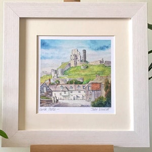 May include: Watercolor painting of Corfe Castle, framed in a white wooden frame. The artwork depicts the castle atop a green hill, with a village in the foreground. The sky is blue with wispy clouds. The painting is signed by the artist.