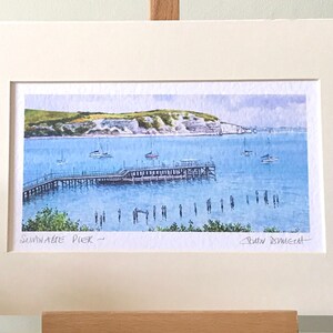 May include: A watercolor painting of a pier extending into the water with a white cliff in the background. The pier is surrounded by boats and people are standing on the pier and in the water. The painting is titled "Sandgate Pier" and signed by the artist "David Dowsett".