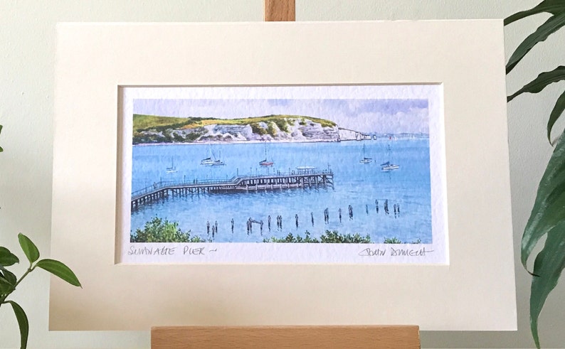 May include: A watercolor painting of a pier extending into the water with a white cliff in the background. The pier is surrounded by boats and people are standing on the pier and in the water. The painting is titled "Sandgate Pier" and signed by the artist "David Dowsett".