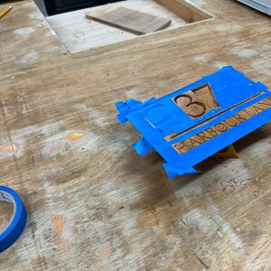 May include: A wooden sign with the number "37" and the text "HARBOUR LANE" is secured with blue painter's tape. The sign is made of light brown wood and is on a wooden surface.