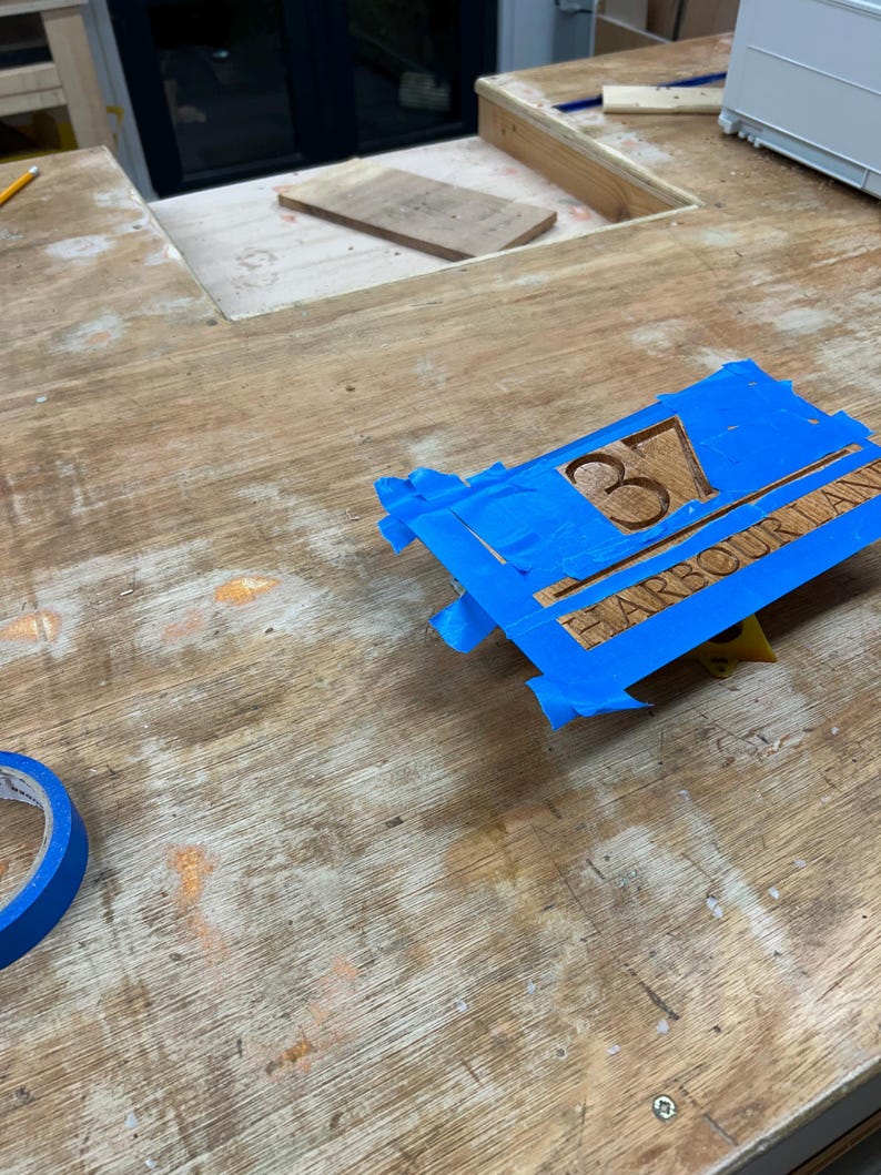 May include: A wooden sign with the number "37" and the text "HARBOUR LANE" is secured with blue painter's tape. The sign is made of light brown wood and is on a wooden surface.