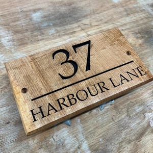 May include: A rectangular wooden sign with the number "37" above the words "HARBOUR LANE" in black lettering. The sign is made of light brown wood with a visible wood grain. Two small holes are drilled on either side of the number.