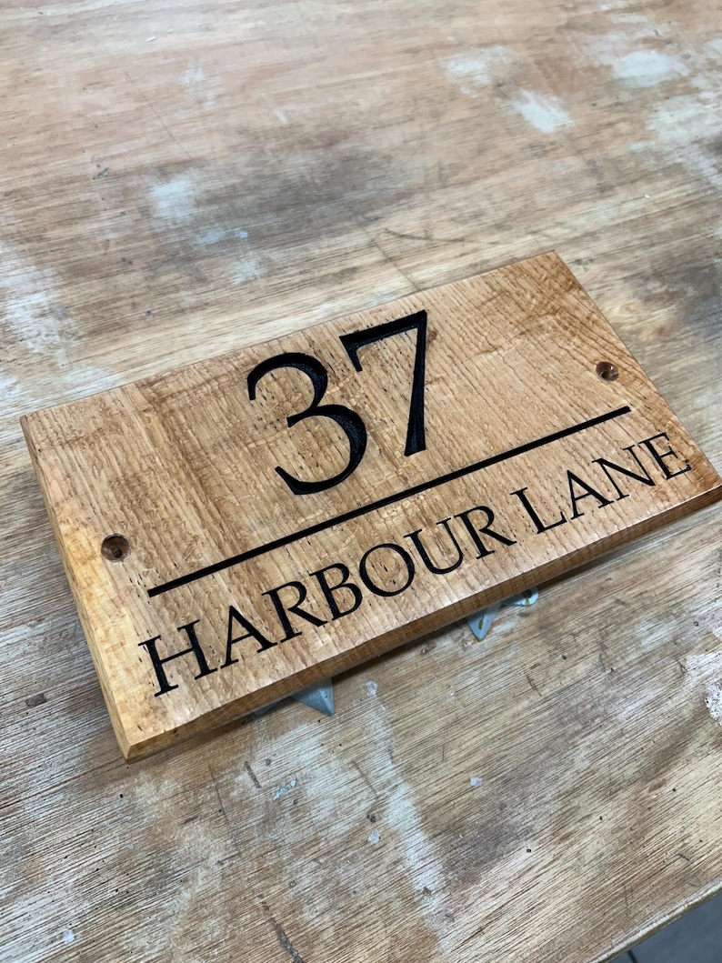May include: A rectangular wooden sign with the number "37" above the words "HARBOUR LANE" in black lettering. The sign is made of light brown wood with a visible wood grain. Two small holes are drilled on either side of the number.