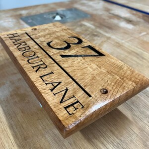 May include: Wooden address sign with the text "HARBOUR LANE" and the number "37" in black. The sign is rectangular, with a natural wood grain finish and two pre-drilled holes for mounting. The sign is a housewarming gift.