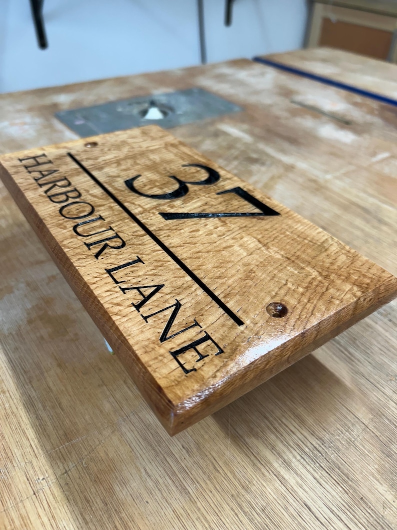 May include: Wooden address sign with the text "HARBOUR LANE" and the number "37" in black. The sign is rectangular, with a natural wood grain finish and two pre-drilled holes for mounting. The sign is a housewarming gift.