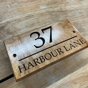 May include: Wooden address sign with the number 37 and the text "HARBOUR LANE" in black lettering. The sign is rectangular with rounded corners and two pre-drilled holes. The wood has a light brown color with a natural grain.