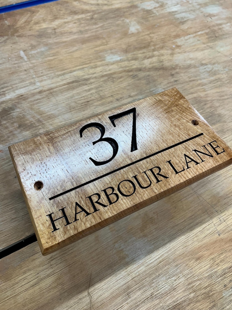 May include: Wooden address sign with the number 37 and the text "HARBOUR LANE" in black lettering. The sign is rectangular with rounded corners and two pre-drilled holes. The wood has a light brown color with a natural grain.