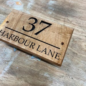May include: A rectangular wooden address sign with the number "37" in bold black font. Below the number, the text "HARBOUR LANE" is also in black. The sign has a natural wood grain finish and two pre-drilled holes.