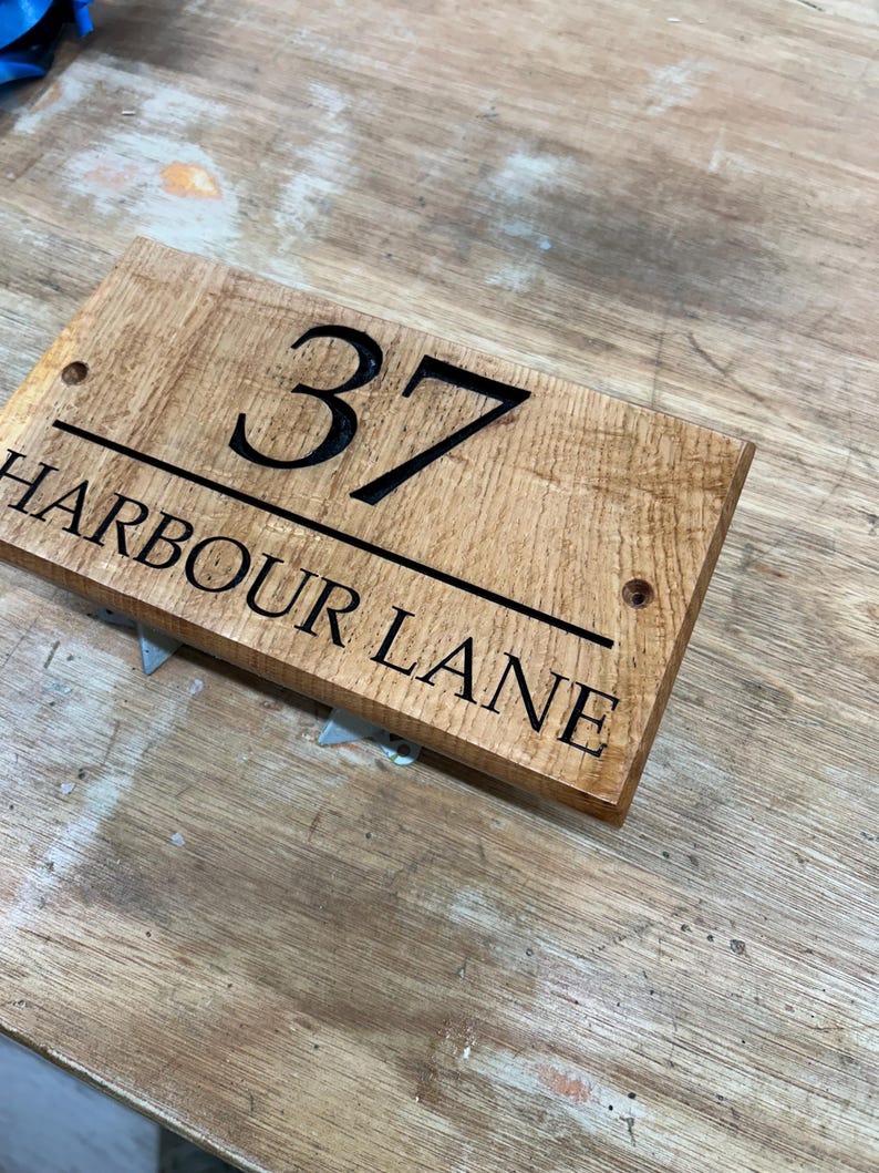 May include: A rectangular wooden address sign with the number "37" in bold black font. Below the number, the text "HARBOUR LANE" is also in black. The sign has a natural wood grain finish and two pre-drilled holes.