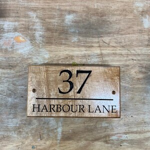 May include: Wooden address sign with the number 37 and the words "HARBOUR LANE" in black lettering. The sign is rectangular and has a natural wood grain finish. Two small screws are visible.