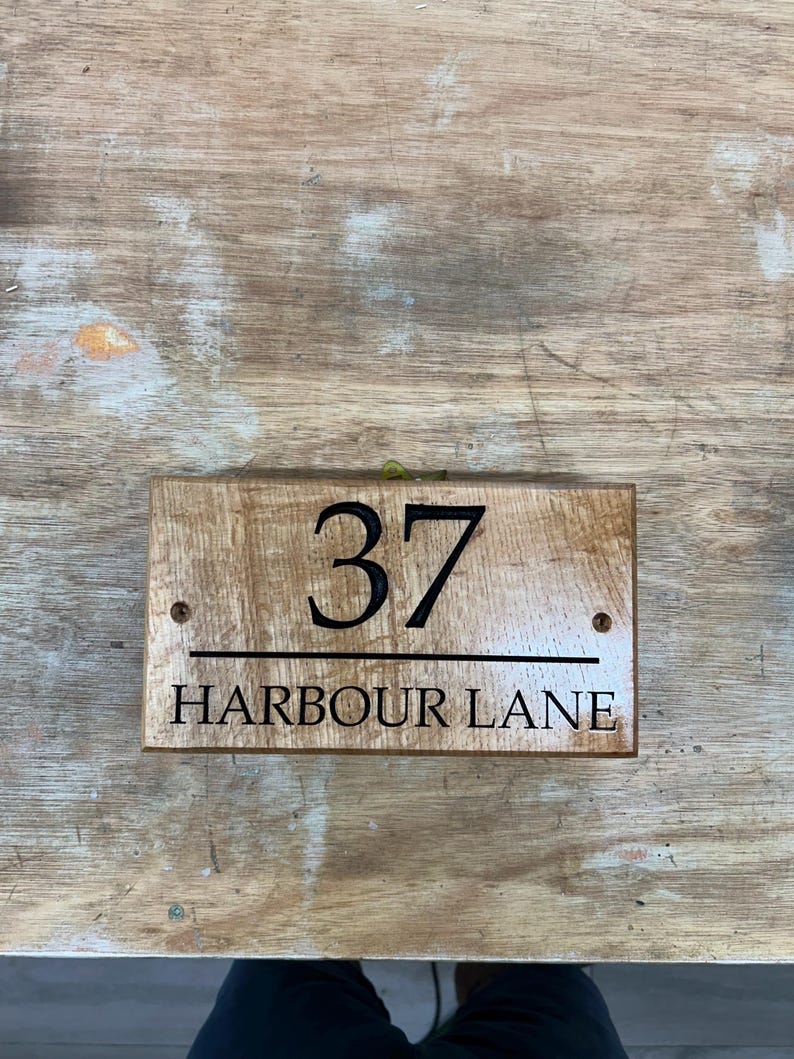 May include: Wooden address sign with the number 37 and the words "HARBOUR LANE" in black lettering. The sign is rectangular and has a natural wood grain finish. Two small screws are visible.