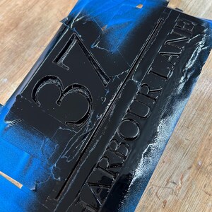 May include: Black rectangular sign with the address "37 HARBOUR LANE" in raised lettering. The sign is partially covered with blue painter's tape and sits on a wooden surface. The numbers and letters are painted black.