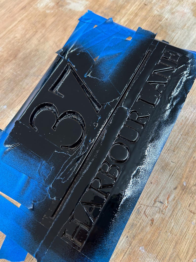 May include: Black rectangular sign with the address "37 HARBOUR LANE" in raised lettering. The sign is partially covered with blue painter's tape and sits on a wooden surface. The numbers and letters are painted black.