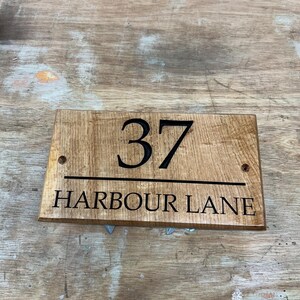 May include: Wooden address sign with the number "37" and the words "HARBOUR LANE" in black lettering. The sign is rectangular with rounded corners and two pre-drilled holes for mounting. The wood has a natural, light brown finish.