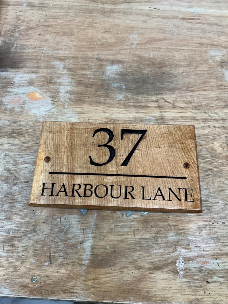 May include: Wooden address sign with the number "37" and the words "HARBOUR LANE" in black lettering. The sign is rectangular with rounded corners and two pre-drilled holes for mounting. The wood has a natural, light brown finish.