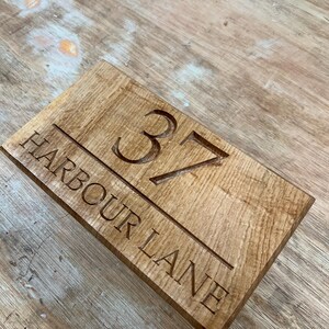 May include: A rectangular wooden sign with the engraved text "37 HARBOUR LANE". The sign is made of light brown wood with visible grain and a carved design. The numbers and letters are deeply carved.
