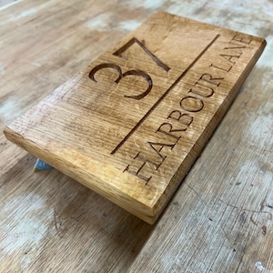May include: A rectangular wooden sign with the engraved number "37" and the words "HARBOUR LANE" in a serif font. The sign is made of light brown wood with a visible wood grain.