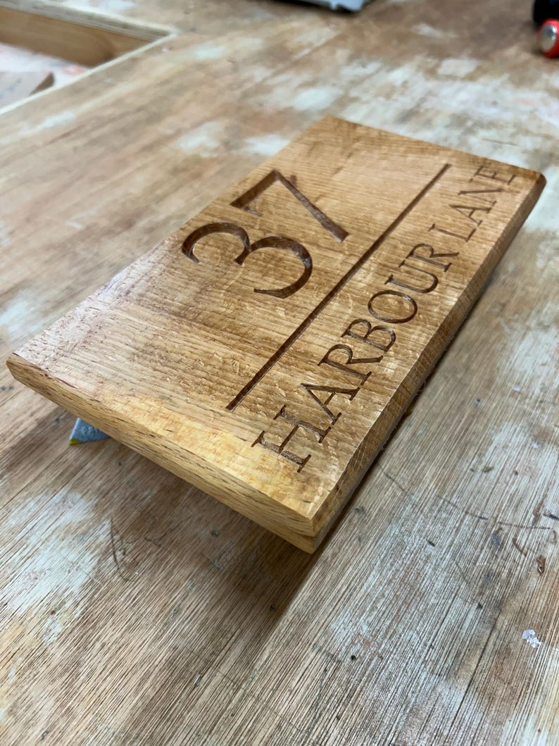 May include: A rectangular wooden sign with the engraved number "37" and the words "HARBOUR LANE" in a serif font. The sign is made of light brown wood with a visible wood grain.