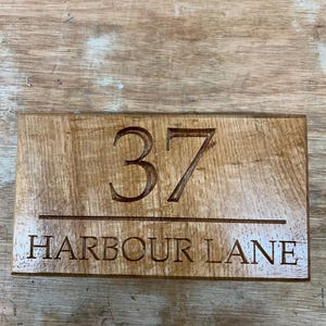 May include: A rectangular wooden sign with the number "37" carved into the wood above the words "HARBOUR LANE". The sign is made of light brown wood with a visible wood grain.