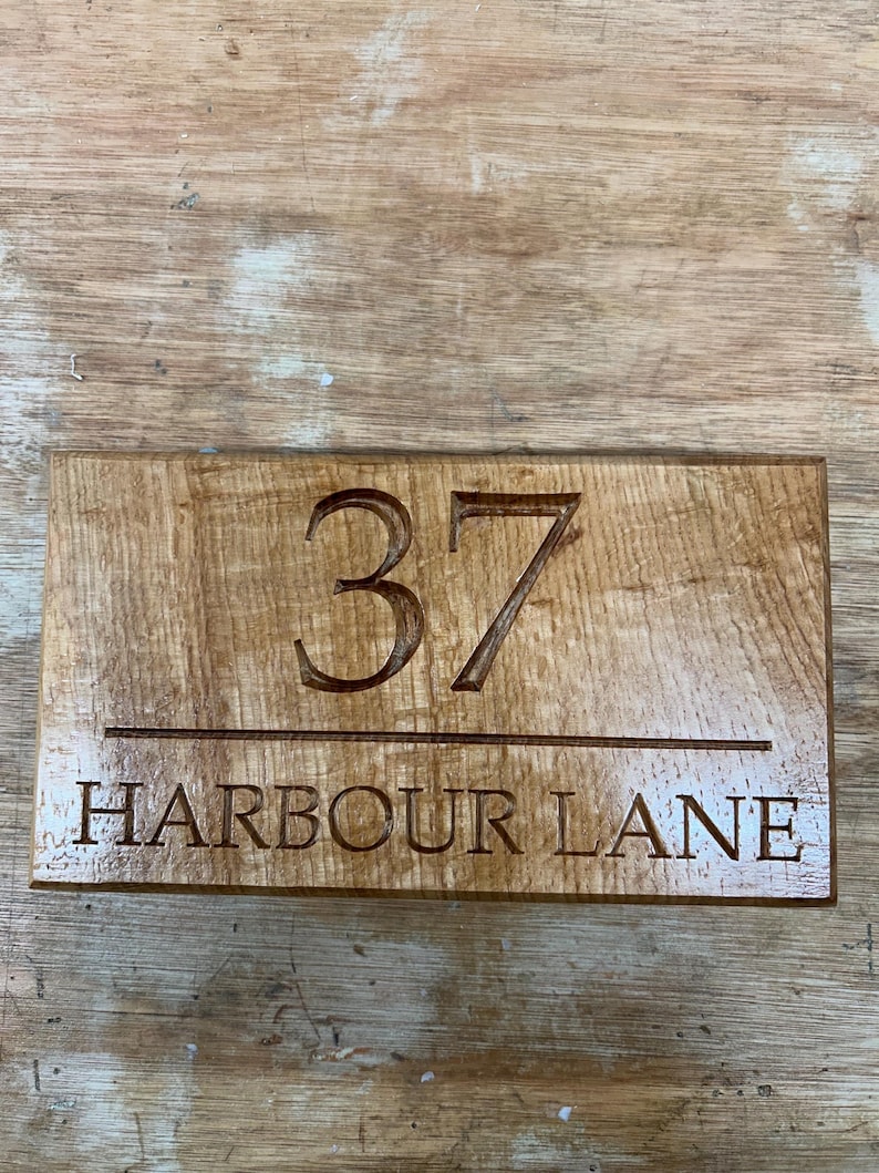 May include: A rectangular wooden sign with the number "37" carved into the wood above the words "HARBOUR LANE". The sign is made of light brown wood with a visible wood grain.