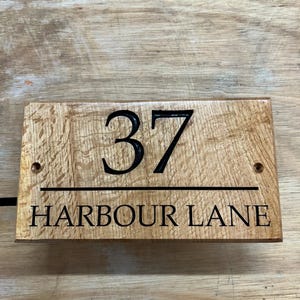 May include: Wooden address sign with the number "37" and the words "HARBOUR LANE" in black lettering. The sign is rectangular and made of light brown wood with a visible wood grain.