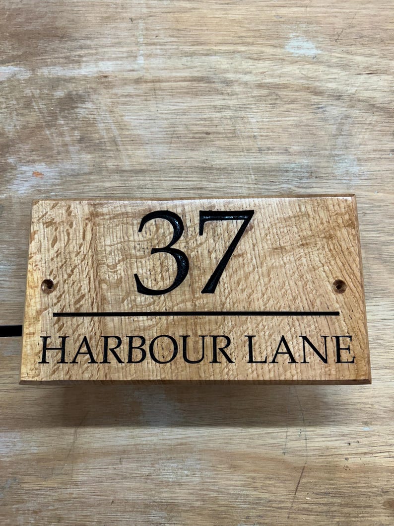 May include: Wooden address sign with the number "37" and the words "HARBOUR LANE" in black lettering. The sign is rectangular and made of light brown wood with a visible wood grain.