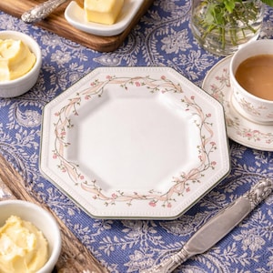 May include: A white octagonal plate with a floral and ribbon design, set on a blue paisley tablecloth. The table setting includes a cup of tea, butter, bread, and a bouquet of pink roses.