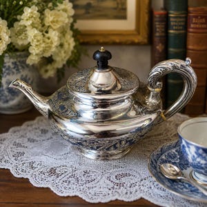 May include: A polished silver teapot featuring intricate embossed floral and paisley patterns across its rounded body, a curved handle, a tapered spout, and a lid topped with a small black knob. The metal surface reflects light.