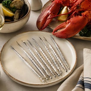 May include: A set of eight silver-toned metal seafood picks arranged in a fan pattern on a round ceramic plate. Each pick features a slender, pointed tip and a textured, decorative handle with a rounded end, designed for extracting meat from shellfish.