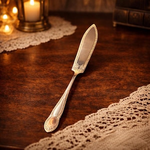 May include: A silver butter knife with an ornate handle and a decorative blade. The knife is set on a dark wooden surface with a lace doily in the background. The knife has a decorative floral design.