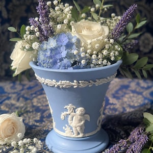 May include: A light blue ceramic vase featuring white relief carvings of cherubs and a laurel leaf border. The vase is filled with a floral arrangement consisting of white roses, blue hydrangea blossoms, purple lavender, and white baby's breath.