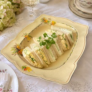 May include: A square, cream-coloured ceramic serving plate with scalloped edges and a delicate floral pattern in yellow and green. The plate holds several triangular egg salad sandwiches garnished with fresh green watercress sprigs.
