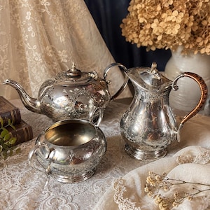 Peut inclure: Un service à thé en argent, comprenant une théière, une crémière et un sucrier, est présenté sur une nappe en dentelle. La théière et la crémière présentent des motifs floraux ornés. La crémière a une poignée marron. L'ensemble est de style vintage.