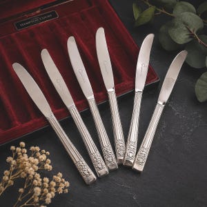 May include: A set of six silver-toned metal knives with smooth blades and handles featuring vertical fluting and a decorative embossed floral motif at the base. Each knife is engraved with the text Hampton Court on the blade surface.