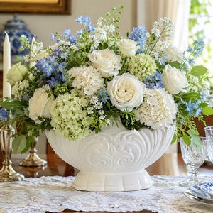 May include: A large floral arrangement featuring white roses, light green hydrangeas, and blue delphinium flowers displayed in a white ceramic bowl with a decorative embossed pattern on the exterior, set on a white lace table runner.