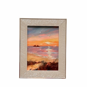 May include: A framed painting depicting a seascape with a sunset over the ocean. The painting features a small island in the distance, a sandy beach, and a few sailboats on the water. The sky is a vibrant orange and pink.